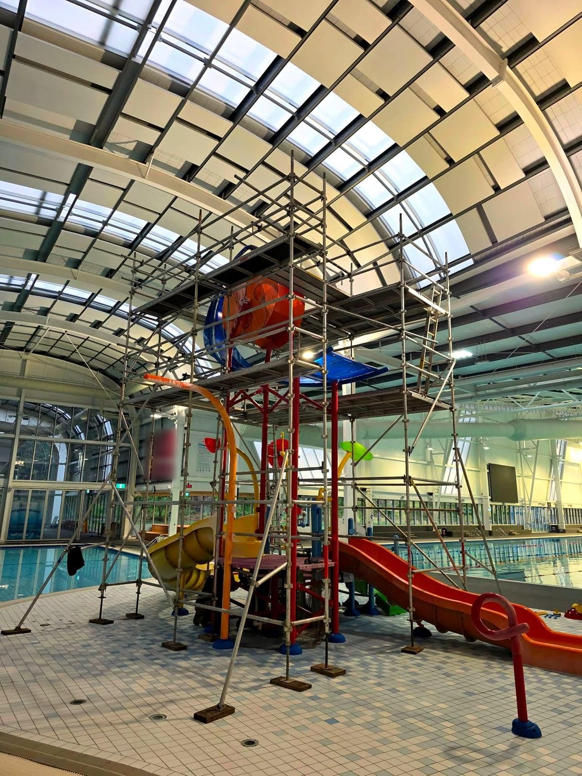 Scaffolding built around an indoor aquatic play structure with slides, under a curved glass roof beside the lap pool